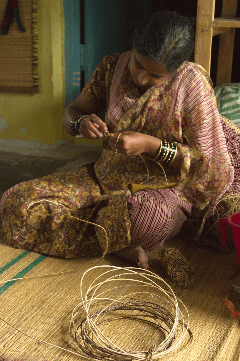 A woman seated on a woven mat, weaving cane by hand, with coils of cane material around her